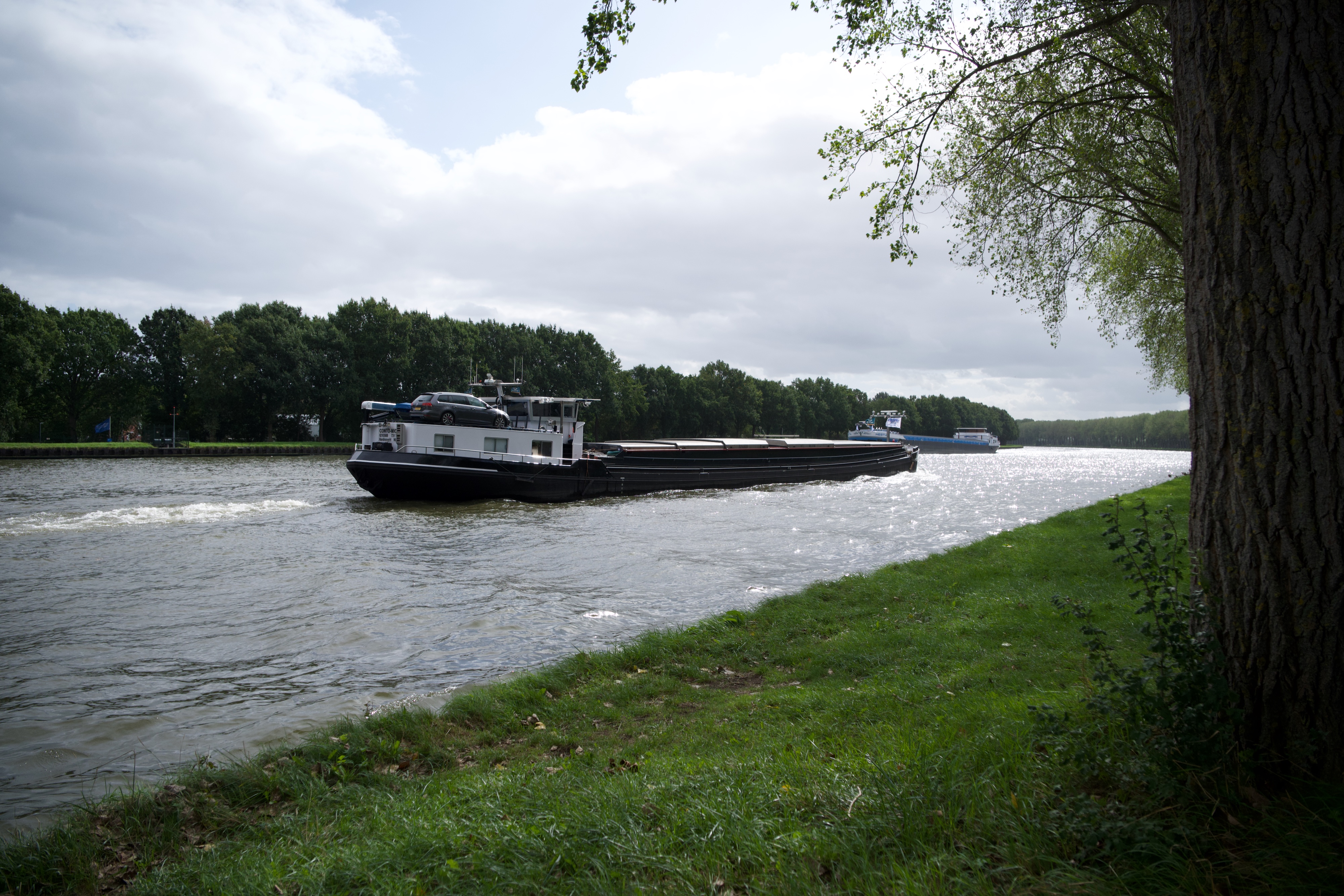 A cargo barge on a canal, trees along the far bank and a cloudy sky.